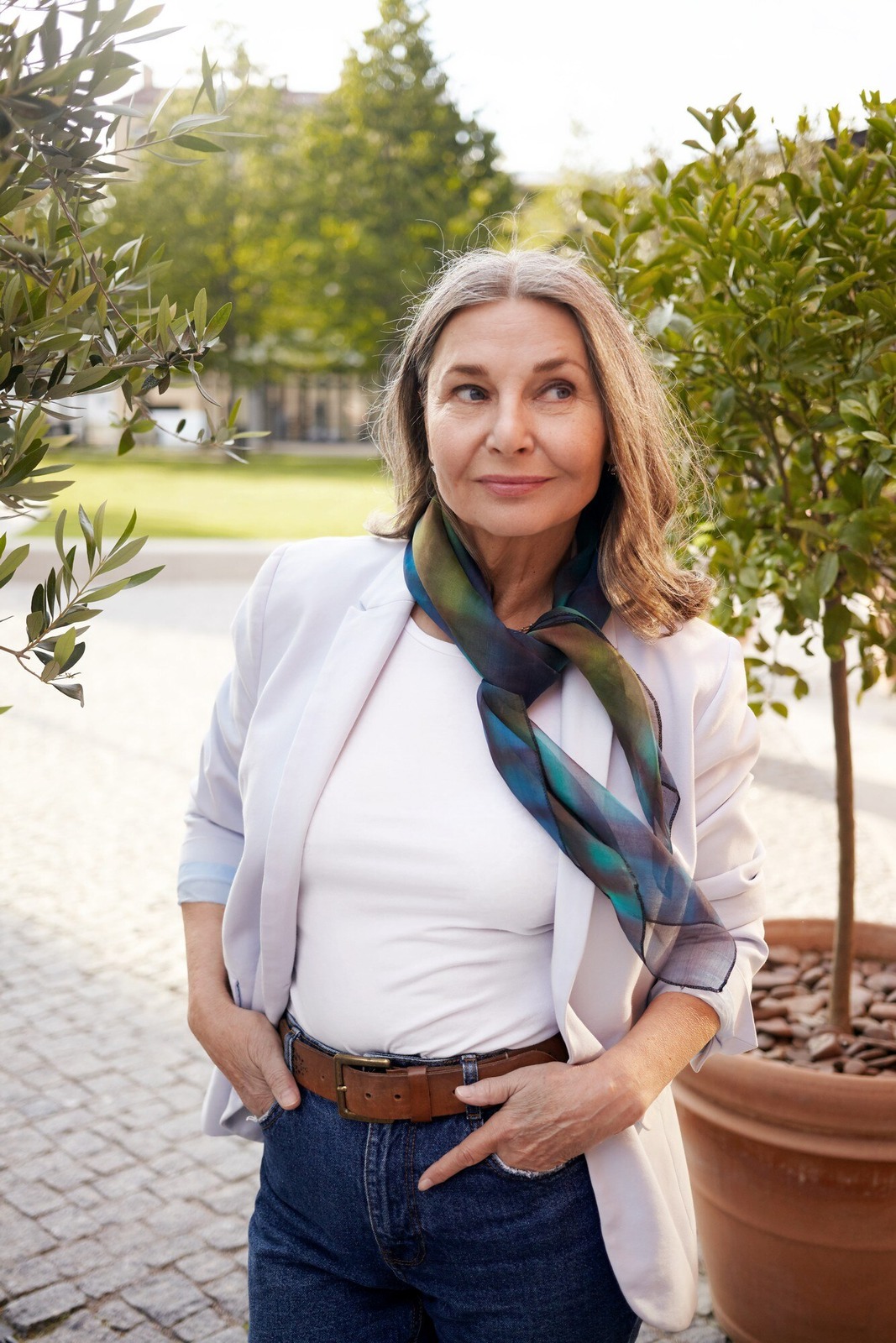 A woman with blonde hair wearing a bright blue dress, sitting on a light-colored sofa in front of large indoor plants, smiling at the camera in an indoor setting with natural light.